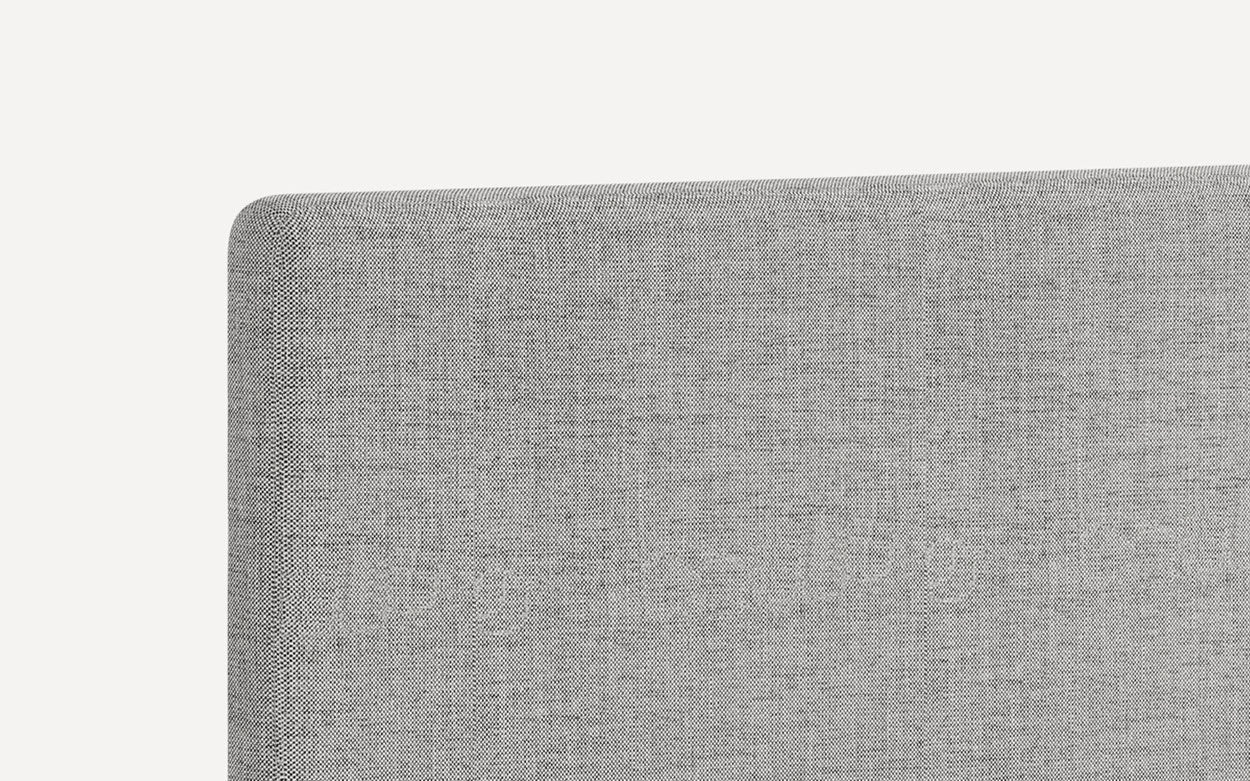Close-up of the top corner of a light gray fabric upholstered headboard with a smooth rounded edge.