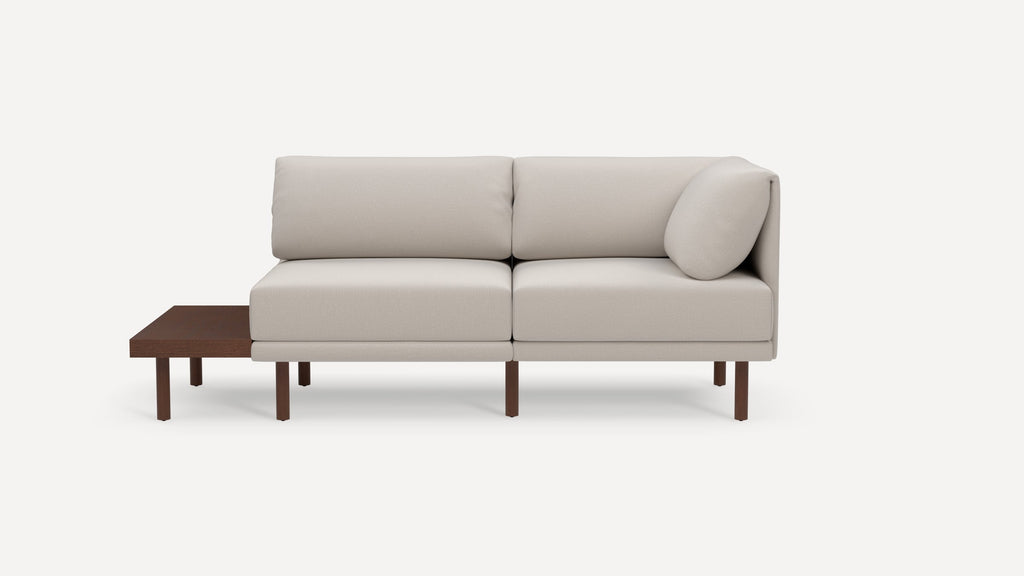Modern beige corner sofa with wooden legs and integrated dark wood side table on left side.