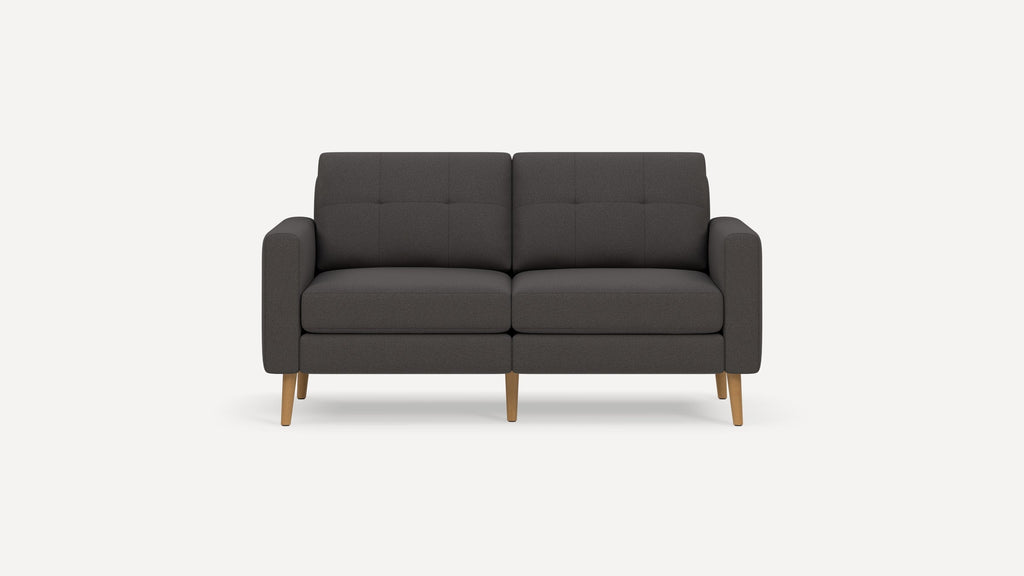 Mid-century modern dark gray upholstered two-seater sofa with tufted back cushions and light wooden legs on white background.