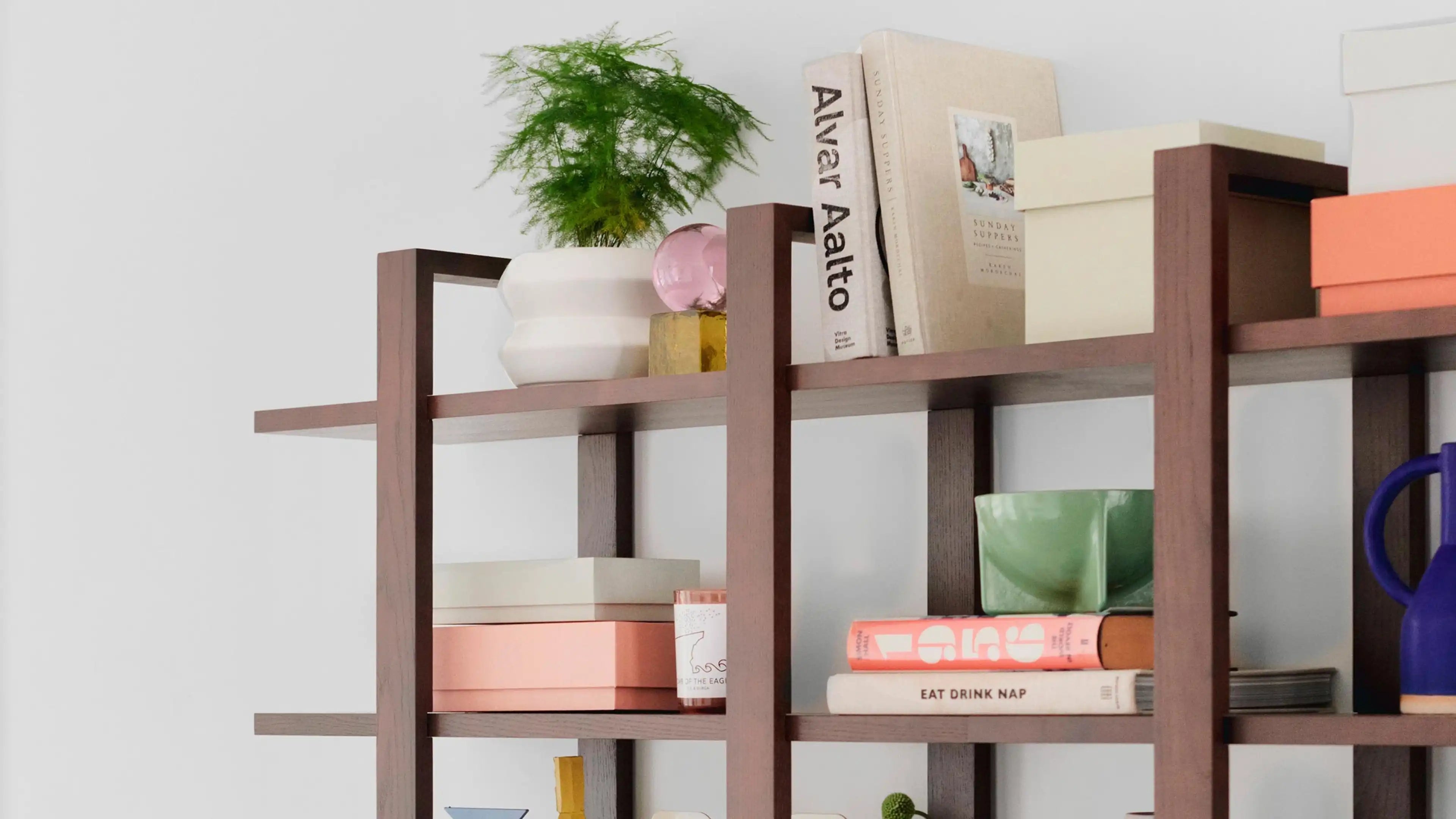 Modern wooden bookshelf with decorative items including books, a white vase with green plant, pastel-colored boxes, and ceramic mugs.