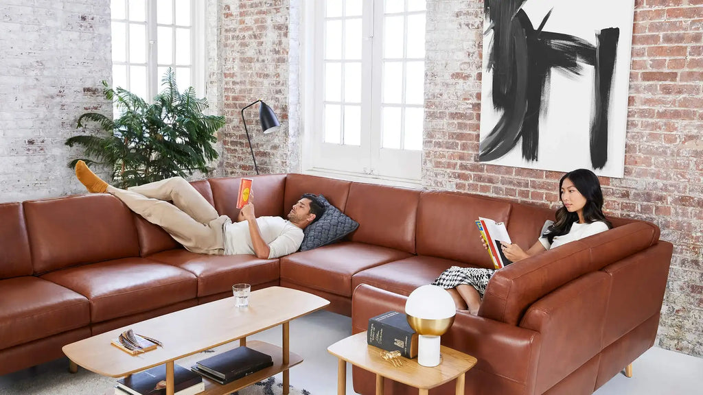 a woman laying on a brown leather sectional sofa in a modern living room with a large wooden coffee table and a large plant on the side table