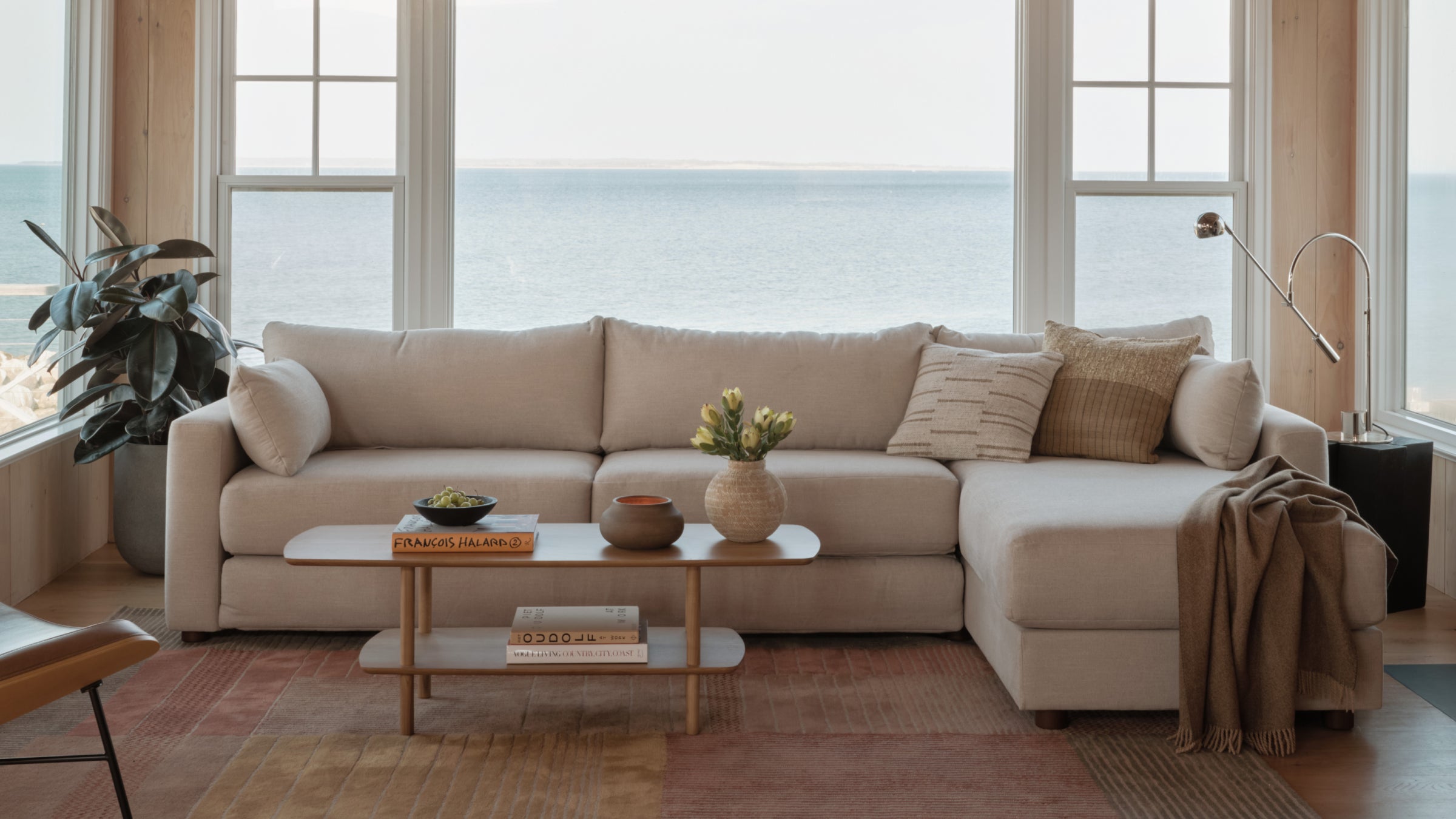 Beige sectional sofa with pillows and throw, wooden coffee table with decor, large windows showing ocean view.