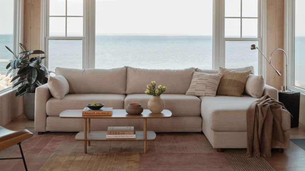 Beige sectional sofa with pillows and throw blanket in a modern living room overlooking a large ocean window view.