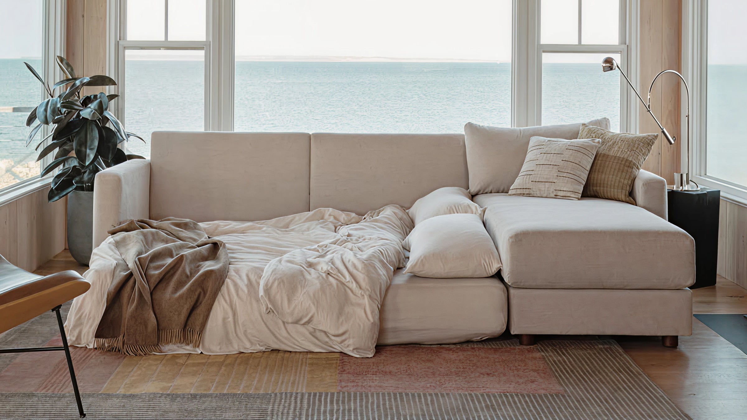 Beige sectional sofa with cushions and cozy throw blanket, placed in a bright room overlooking a sea view.