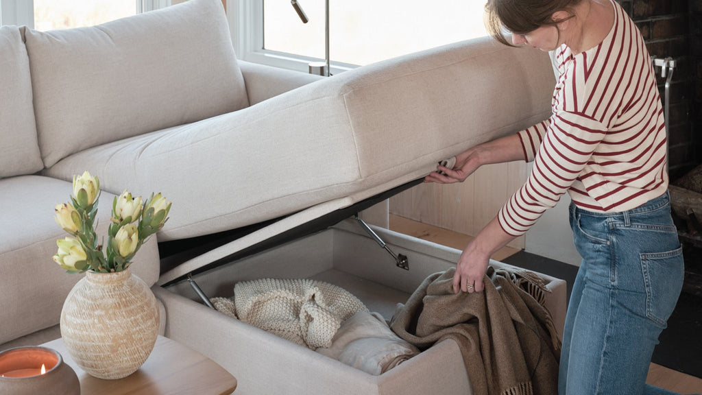Woman lifting beige storage ottoman lid revealing blankets inside, next to a vase with flowers on a wooden table.