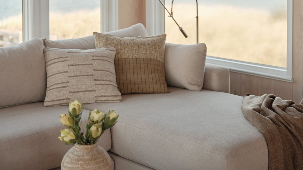 Light beige sectional sofa with three textured neutral-tone pillows, a brown throw blanket, and a decorative vase with flowers.