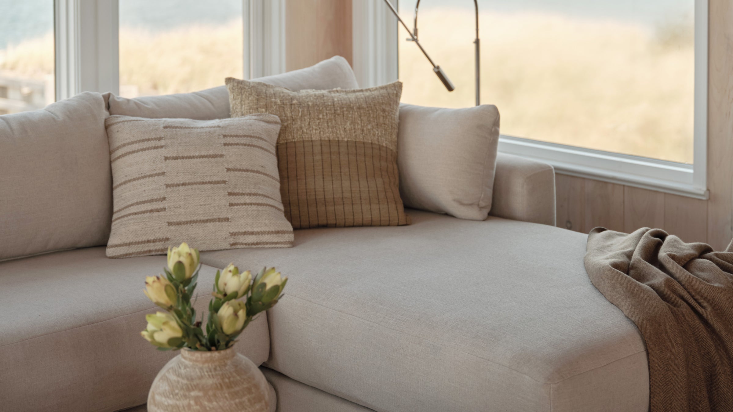 Light beige sectional sofa with textured decorative pillows and a brown throw, set near large windows with natural light.