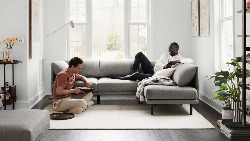 Two men enjoying snacks and drinks on and beside a modern gray sectional sofa in a bright, cozy living room.