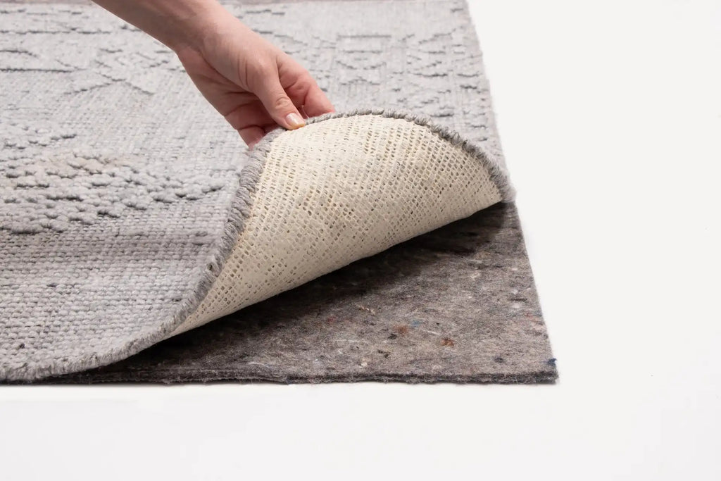 Hand lifting corner of textured gray area rug showing fabric backing and non-slip rug pad underneath on white surface.