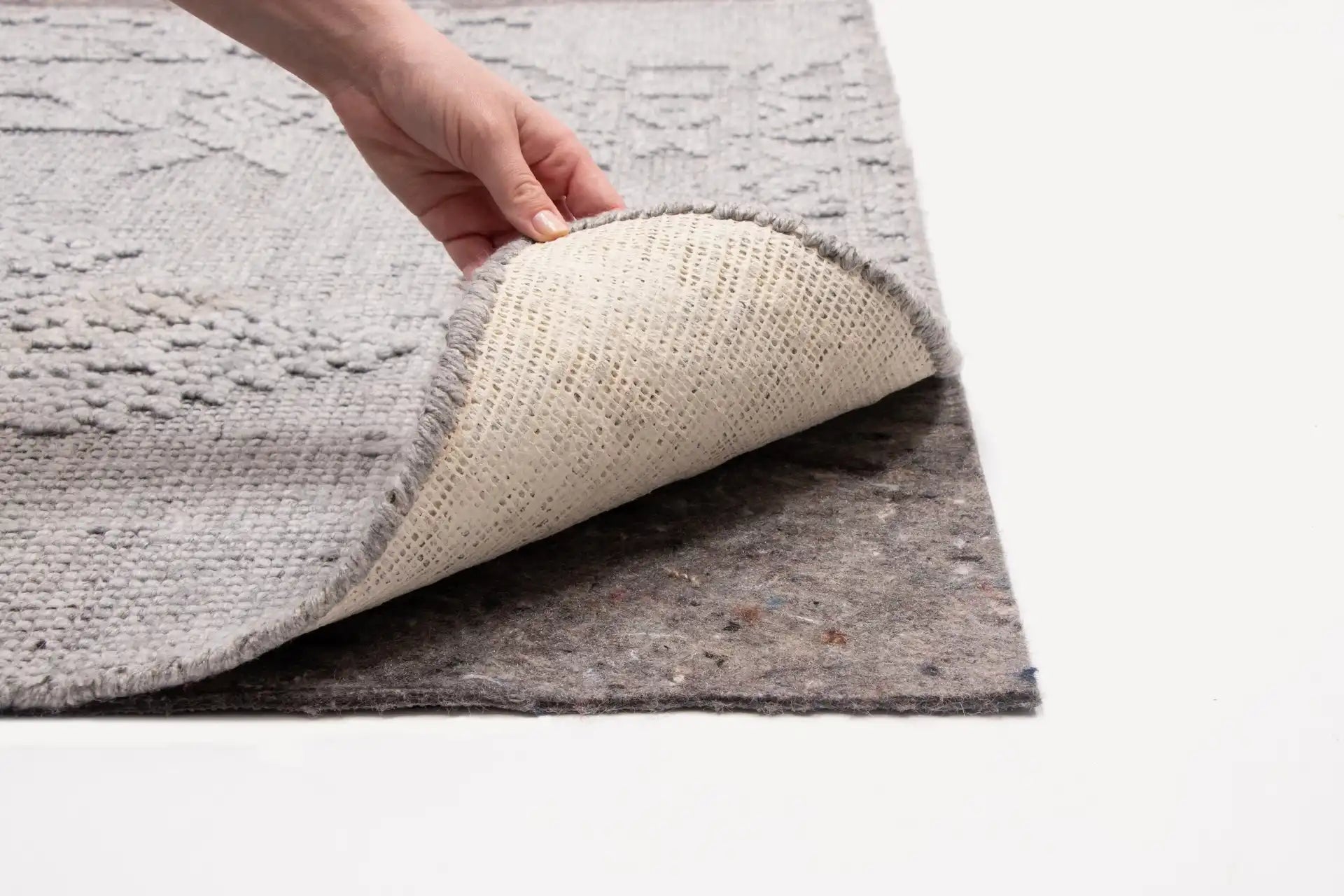 Hand lifting corner of textured gray area rug showing fabric backing and non-slip rug pad underneath on white surface.